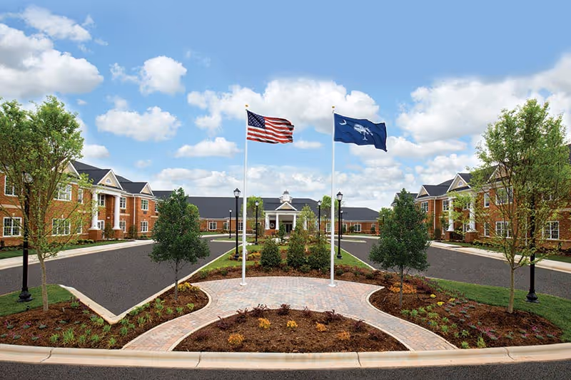 Exterior view of Wellmore of Tega Cay senior living facility showing a circular driveway with landscaped garden in the center, two flagpoles with the American flag and a state flag, surrounded by brick buildings with white-trimmed windows and green trees under a partly cloudy blue sky.