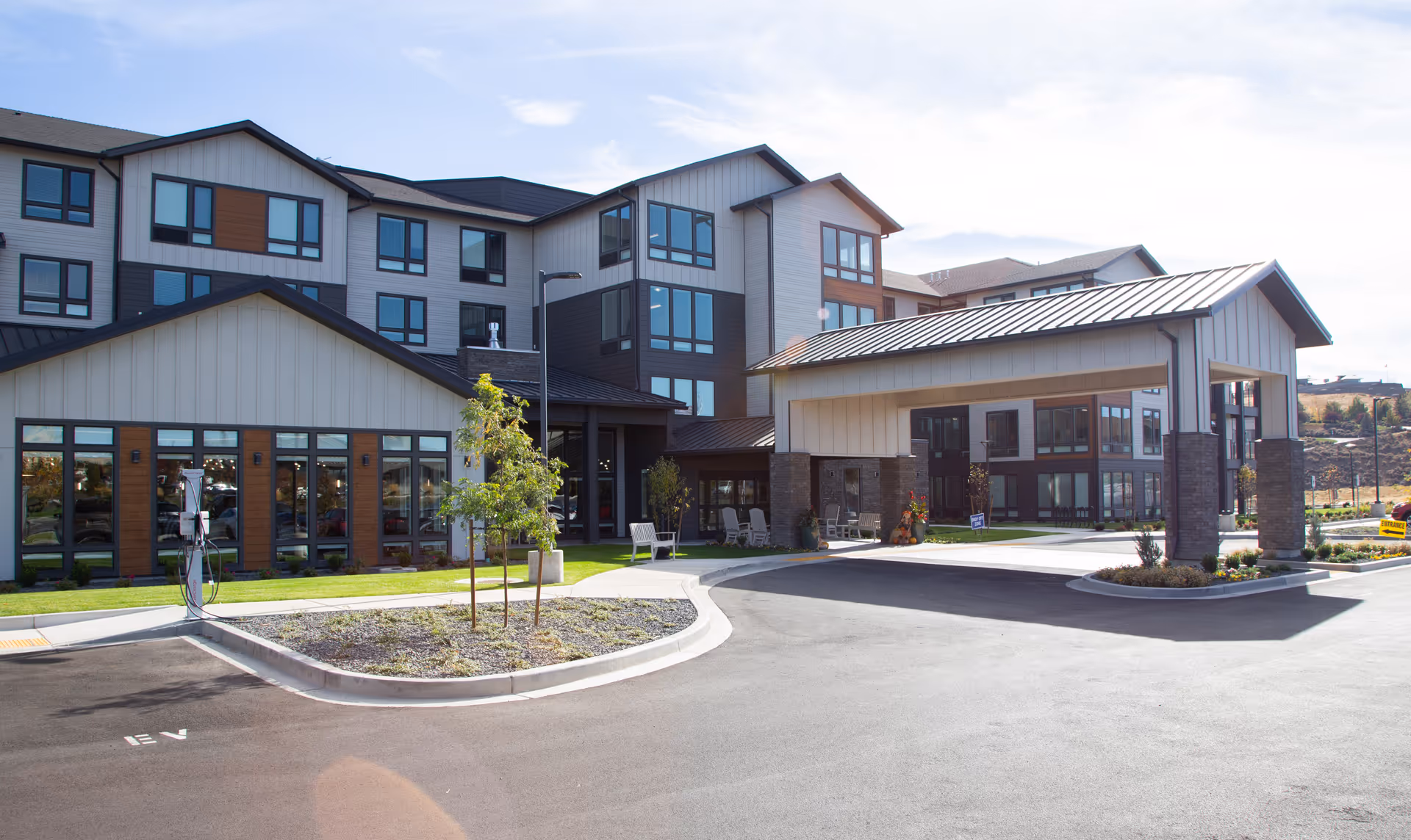Modern multi-story senior living facility exterior with entrance, driveway, and outdoor seating.