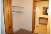 View of a small empty closet with wooden shelves and a hanging rod, adjacent to a bathroom with a sink, mirror, and shower curtain visible.