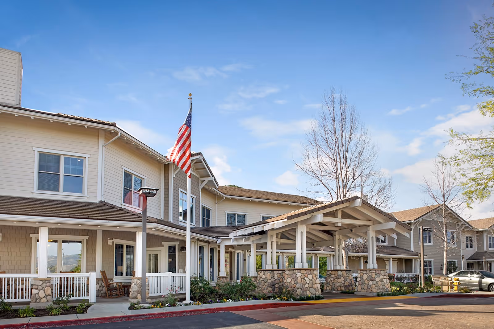 Exterior view of a senior living facility building with beige siding and white trim. The building has a covered entrance supported by stone pillars and an American flag flying on a flagpole in front. There are leafless trees and some greenery around the entrance under a partly cloudy blue sky.