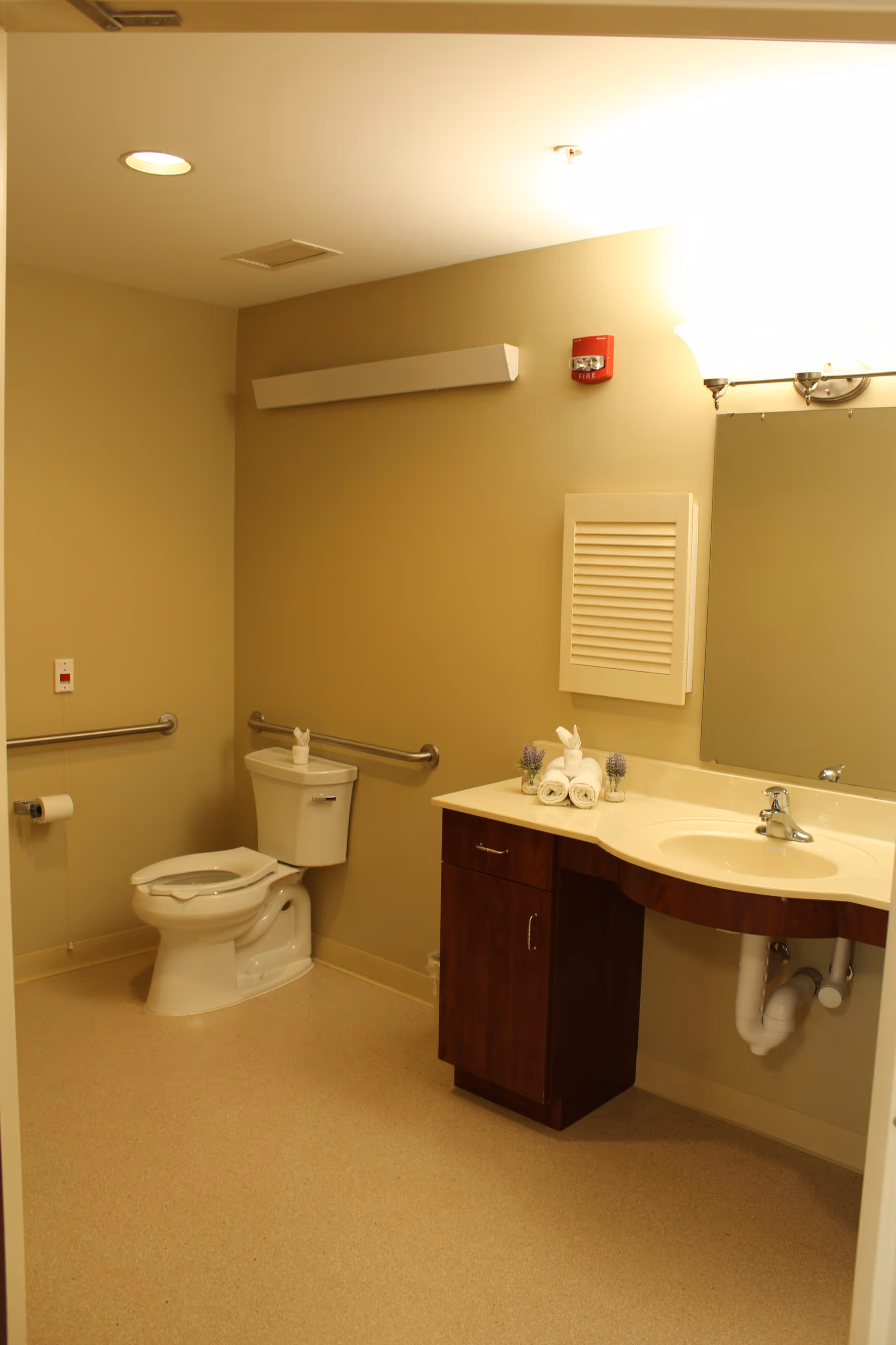 A clean and accessible bathroom with beige walls and flooring. The room features a white toilet with grab bars on the walls for support, a wooden vanity with a cream-colored countertop and an inset sink, a large mirror above the sink, and neatly rolled white towels with small decorative items on the counter.