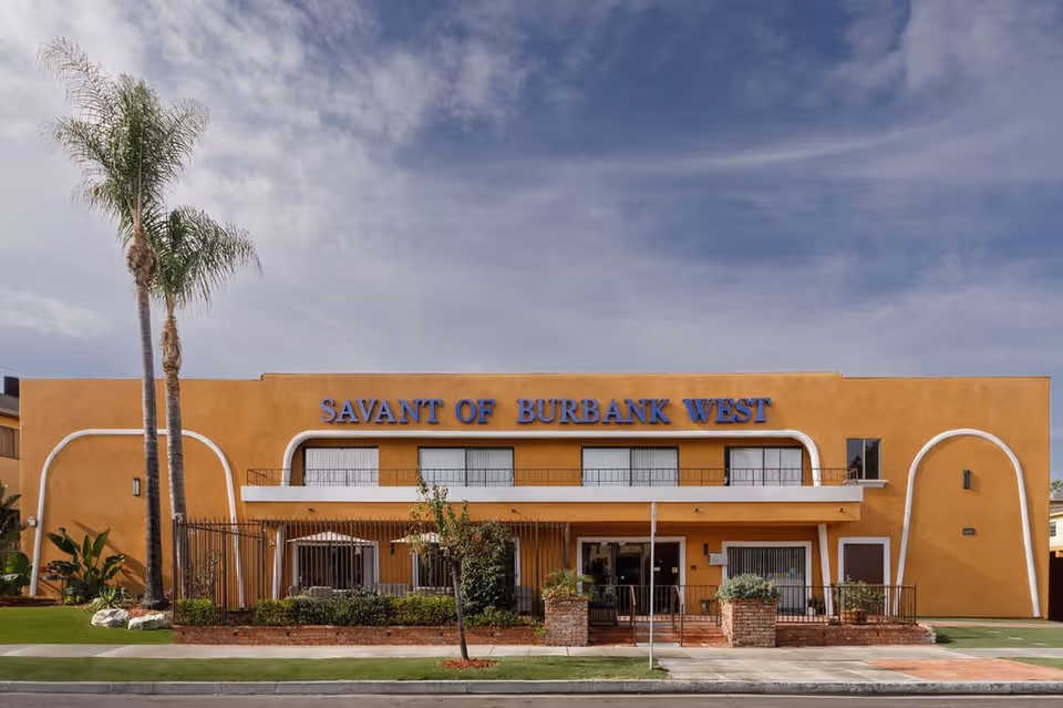 Front exterior of the Savant of Burbank West building with an orange facade, balcony, and palm trees in front.
