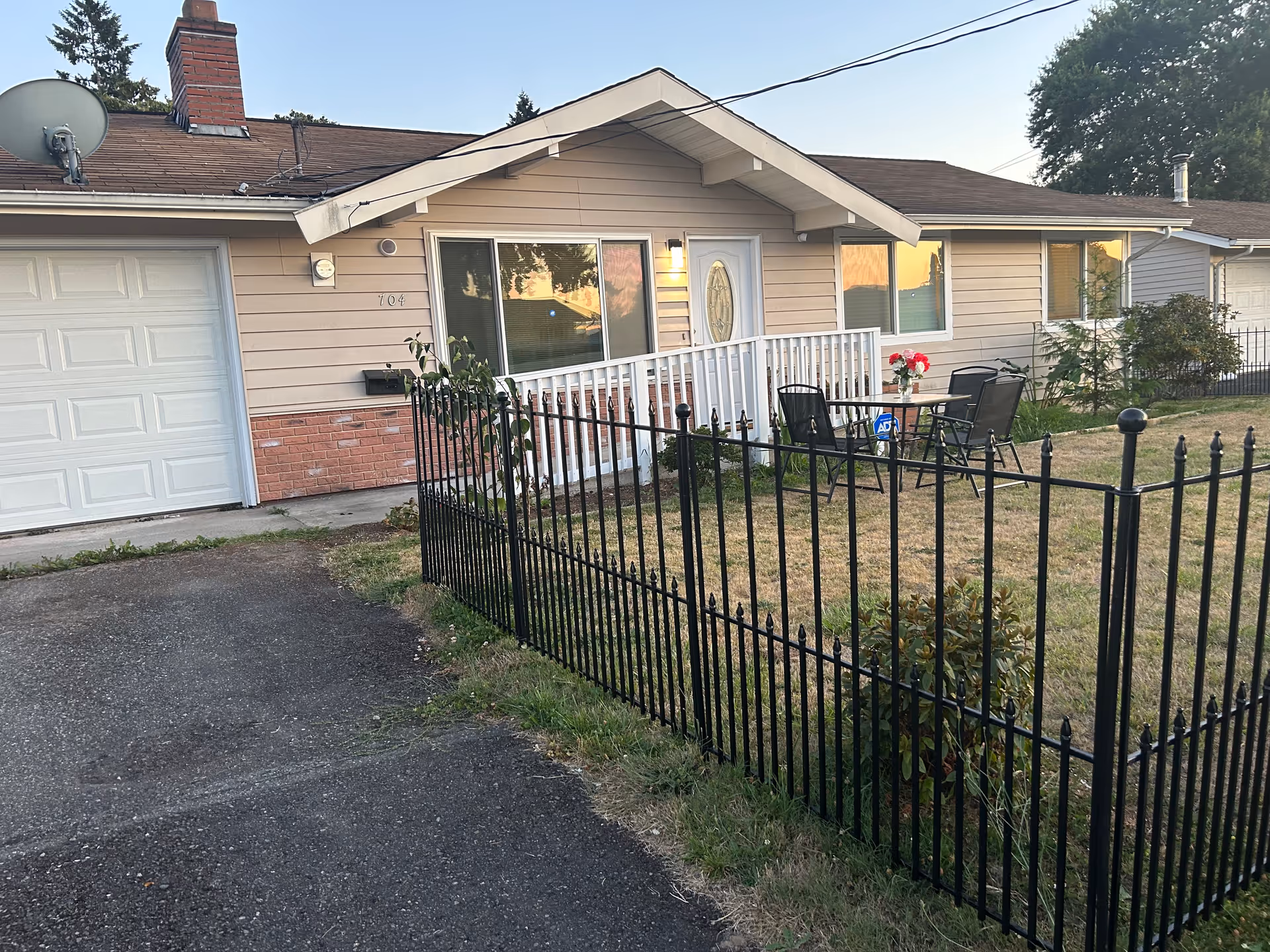 Single-story beige home with white porch railing and outdoor seating behind a black fence.