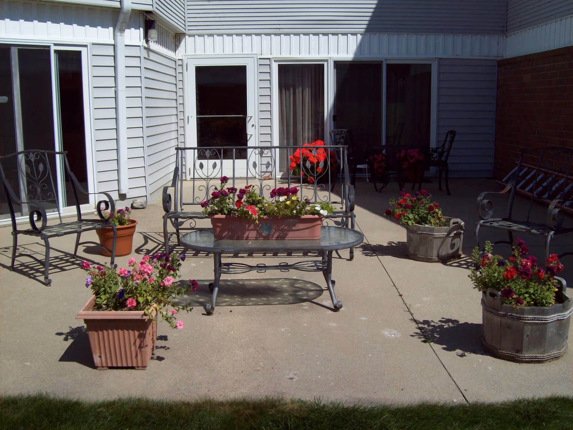 Outdoor patio area with metal chairs and a glass-top table surrounded by several flower pots filled with colorful flowers. The patio is adjacent to a building with white siding and sliding glass doors.