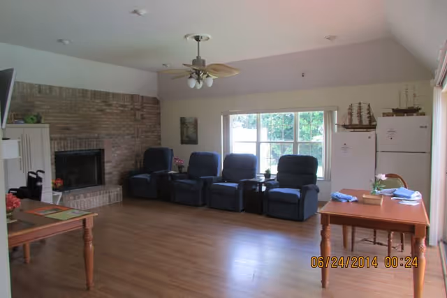 Senior living lounge with recliners, a brick fireplace, and a dining table by a large window.