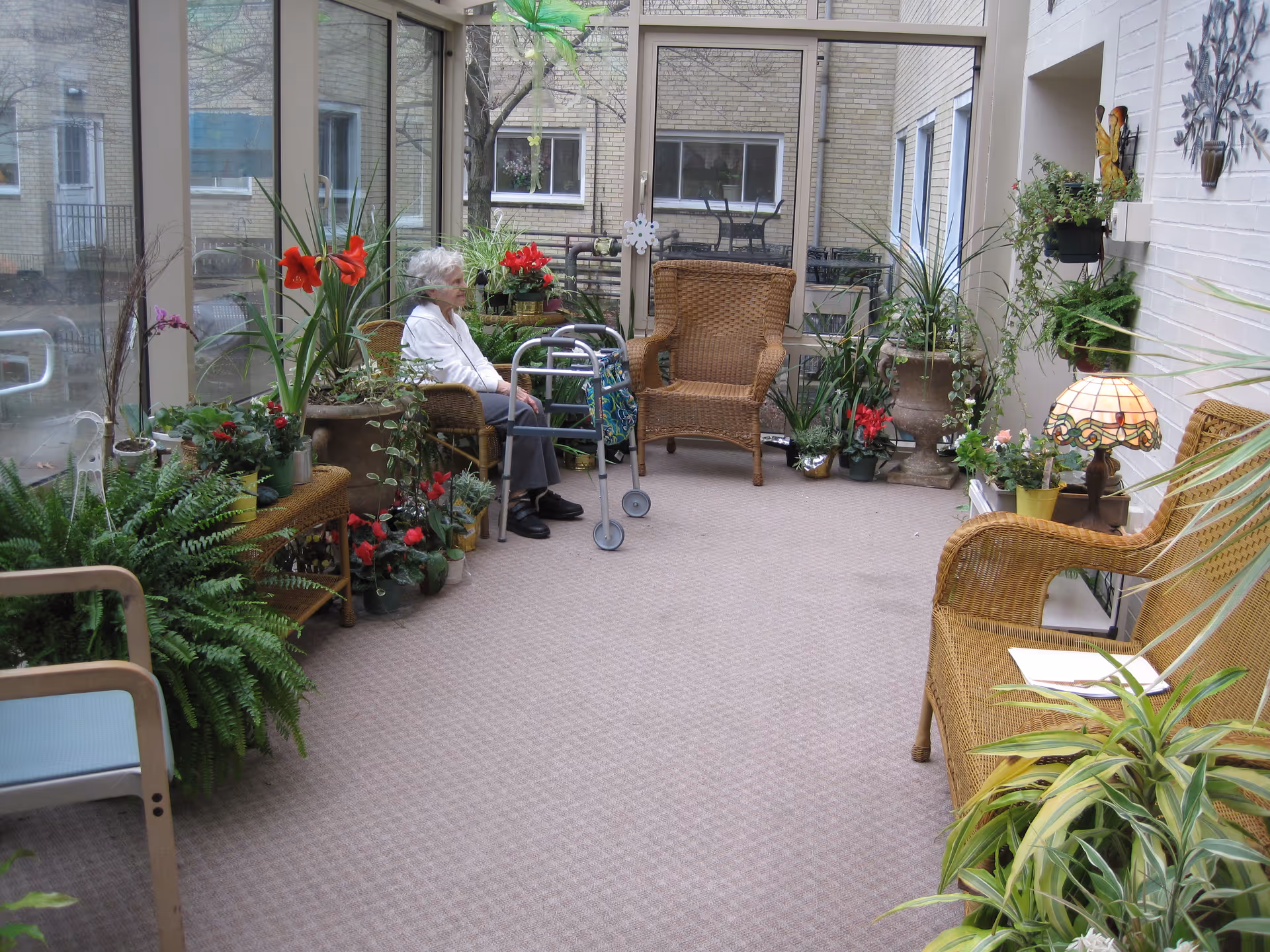 Elderly woman sitting with a walker in a sunroom-style lounge filled with wicker chairs and many potted plants.