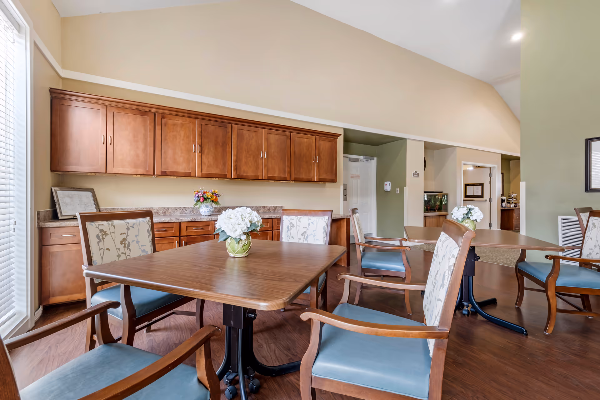 Senior living dining area with wooden tables, chairs, and cabinetry along the wall.