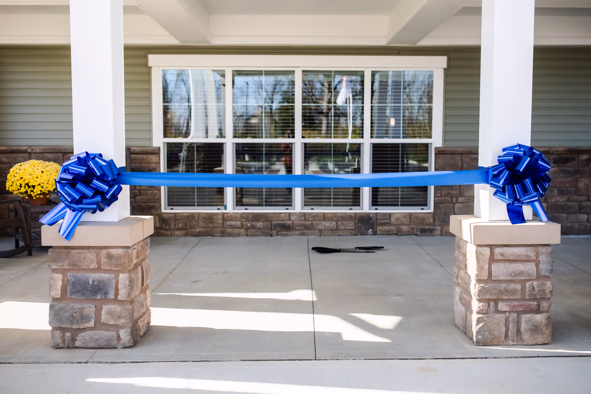 A blue ceremonial ribbon with large bows is stretched between two stone columns across a building's front porch.