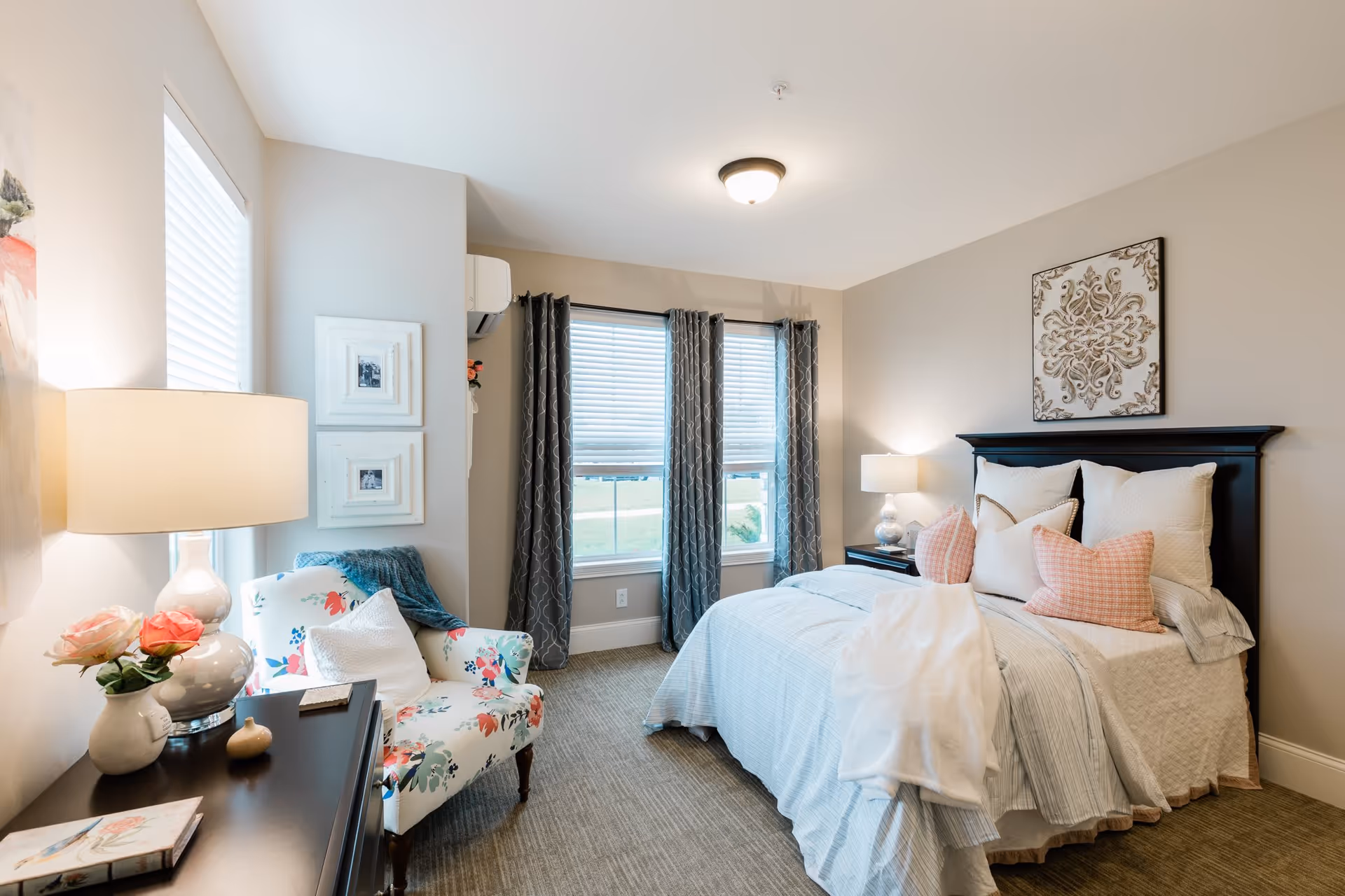 Well-lit bedroom with a made bed, decorative pillows, a floral armchair, bedside tables with lamps, and windows with curtains.