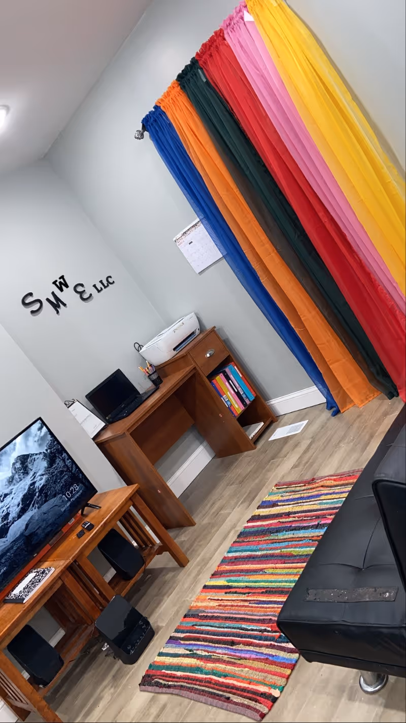 Office/staff area with rainbow fabric streamers, desk, computer, printer, TV, couch, and colorful rug.