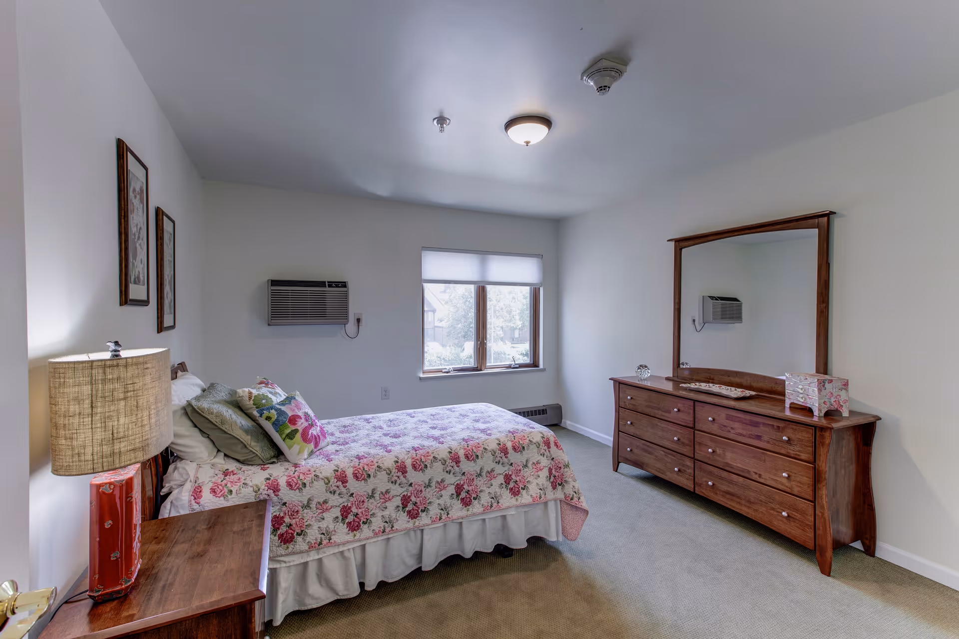 Bright furnished bedroom with a floral bedspread, wooden dresser and mirror, nightstand with lamp, and a window air conditioner.