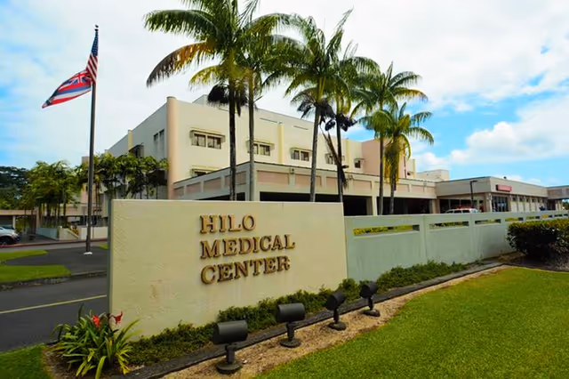 Front view of Hilo Medical Center with a sign, palm trees, and blue sky.