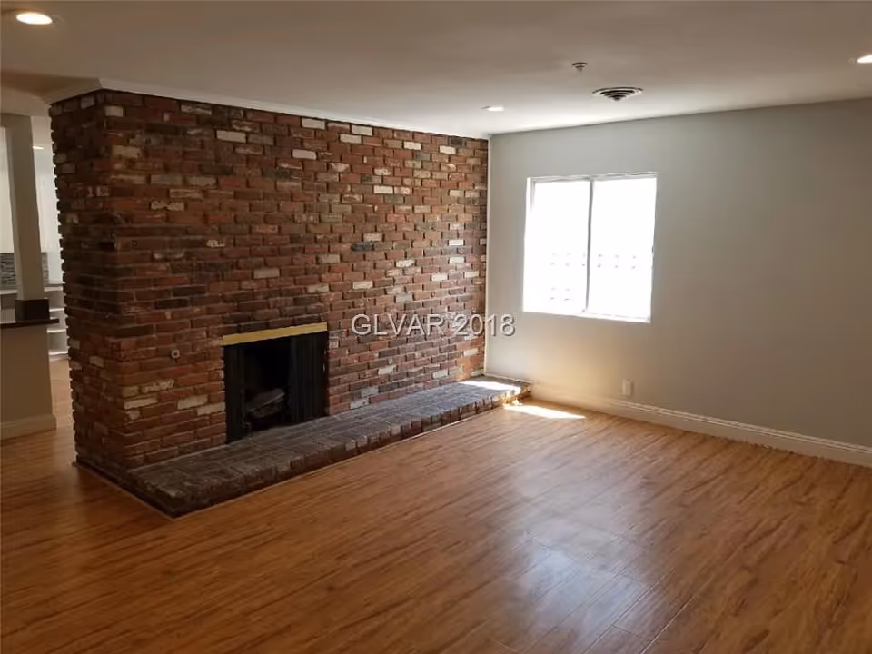 Empty room with wooden flooring, a large brick fireplace on one wall, and a window letting in natural light.