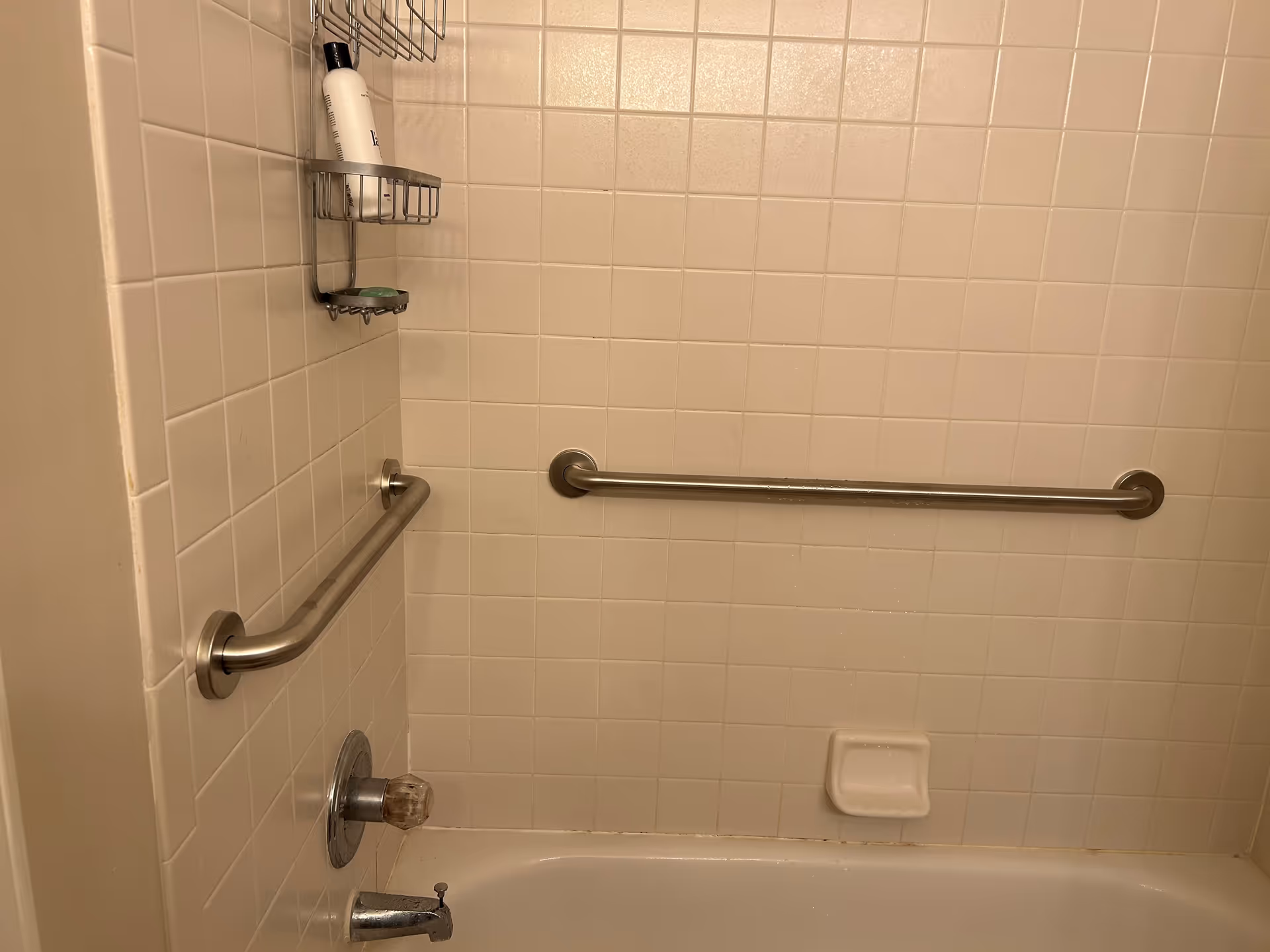 Shower and tub area with wall-mounted grab bars, soap shelf, and tiled walls.