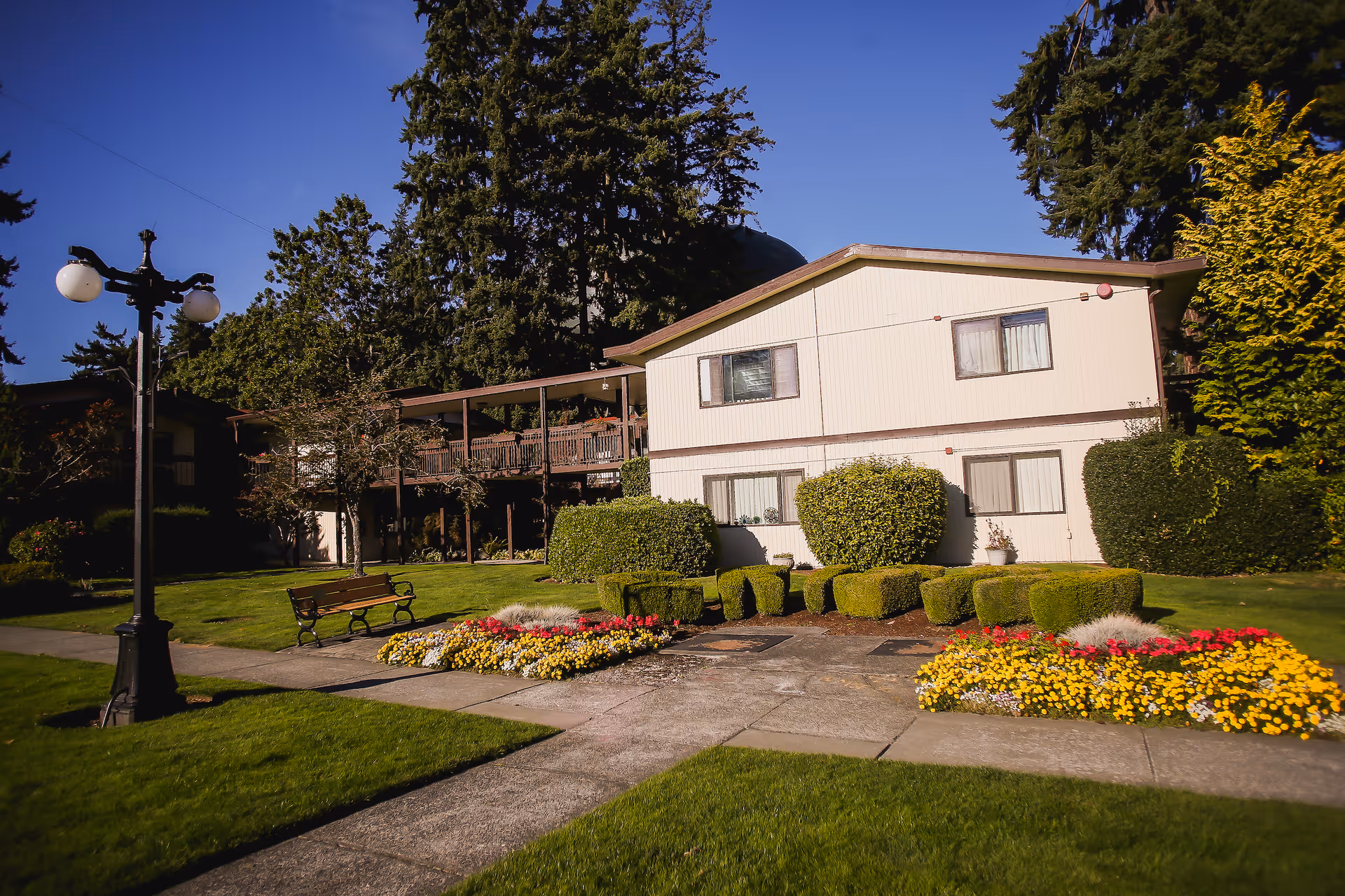 Two-story light-colored building with manicured lawn, flowerbeds, a bench and lamppost under a blue sky.