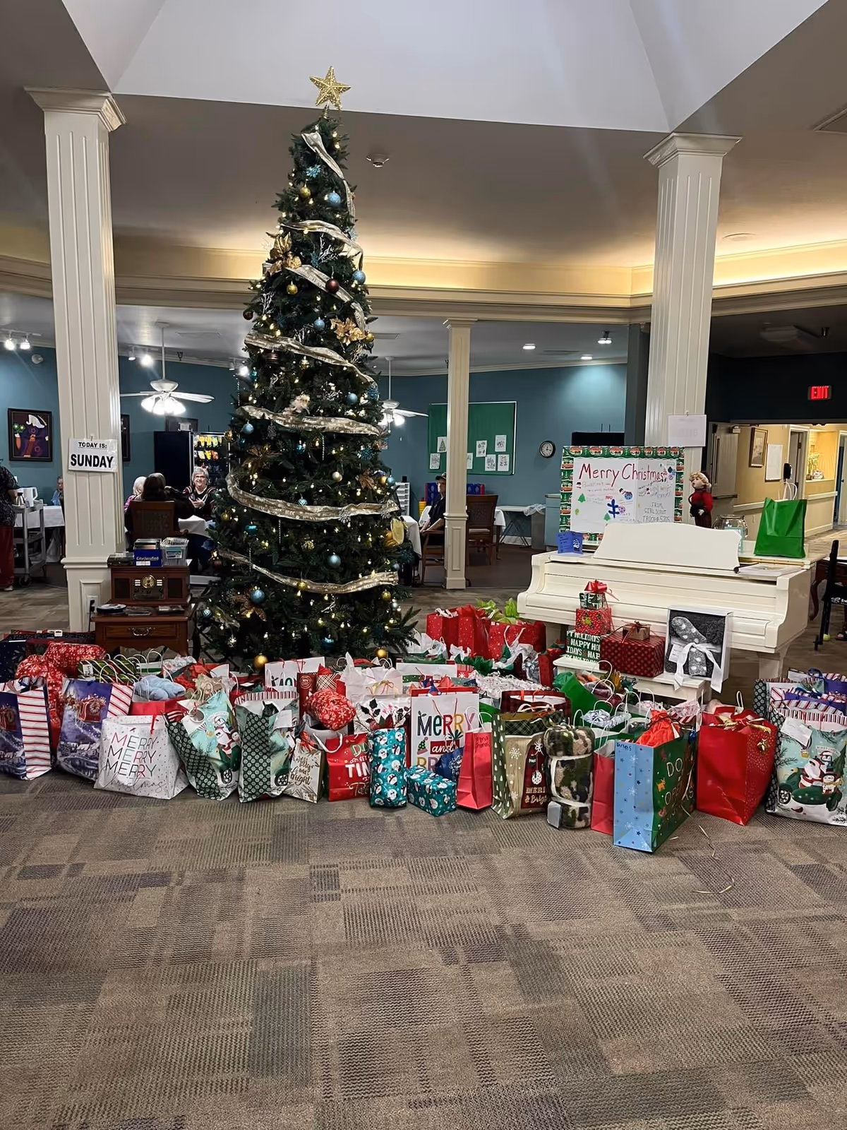 Decorated Christmas tree with wrapped gifts and a white piano in a senior living common area.