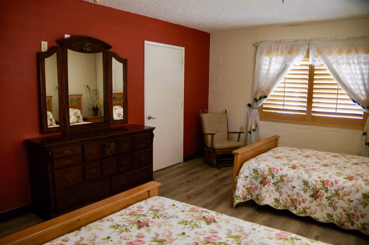 Two beds with floral quilts, dark dresser with mirror, and a chair by the window.