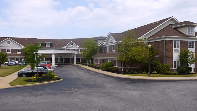 Front driveway and entrance of a two-story brick senior living building with parked cars and landscaping under a partly cloudy sky.