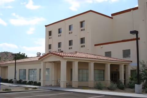 Exterior view of a multi-story assisted living facility with beige walls and a red-tiled roof. The building has several windows and a covered entrance supported by columns. There is a parking area and some landscaping with bushes and small plants around the building.