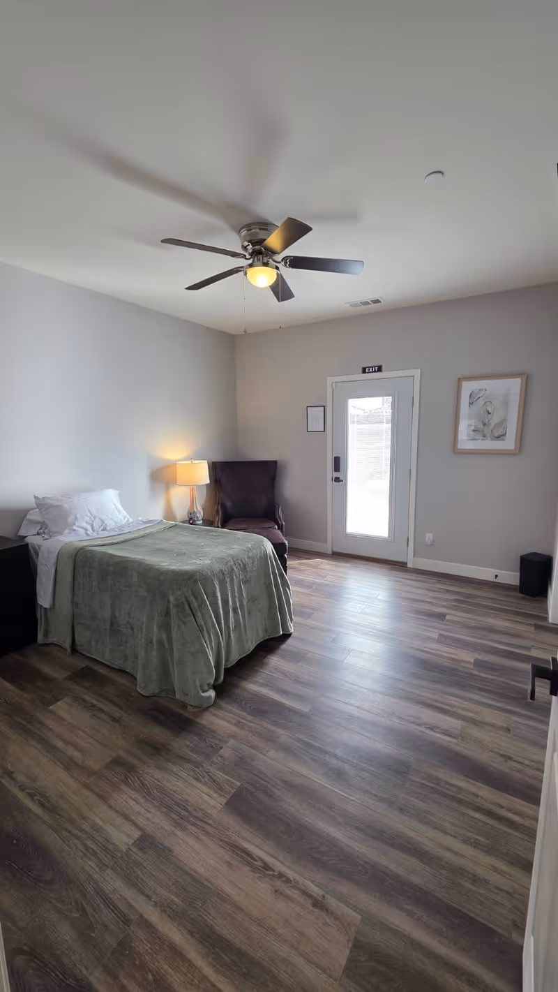 Spacious bedroom in a senior living facility with bed, chair, lamp, and doorway.