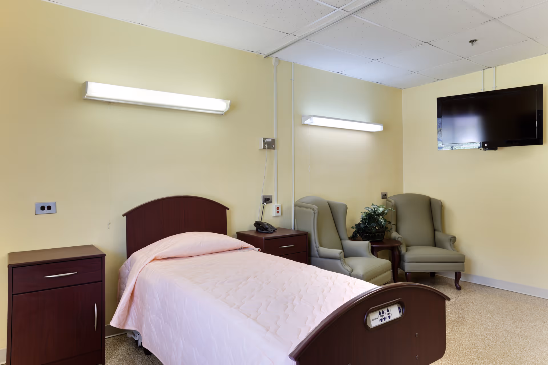 Bedroom in a senior facility with a bed, nightstands, beige chairs, pink quilt, and TV.