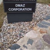 Exterior sign for DMAZ Corporation beside a gravel bed and stone walkway.