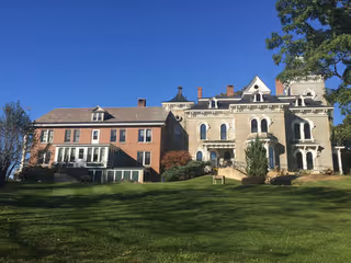 A large historic building with a combination of brick and stone exterior walls, multiple chimneys, and various roof styles under a clear blue sky. The building is set behind a well-maintained grassy lawn with some trees on the sides.