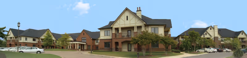 Wide exterior view of Oak Grove Inn, a large senior living facility with multiple connected buildings featuring brick and beige siding, pitched roofs, and several windows. There are trees and parked cars along the driveway under a clear blue sky.