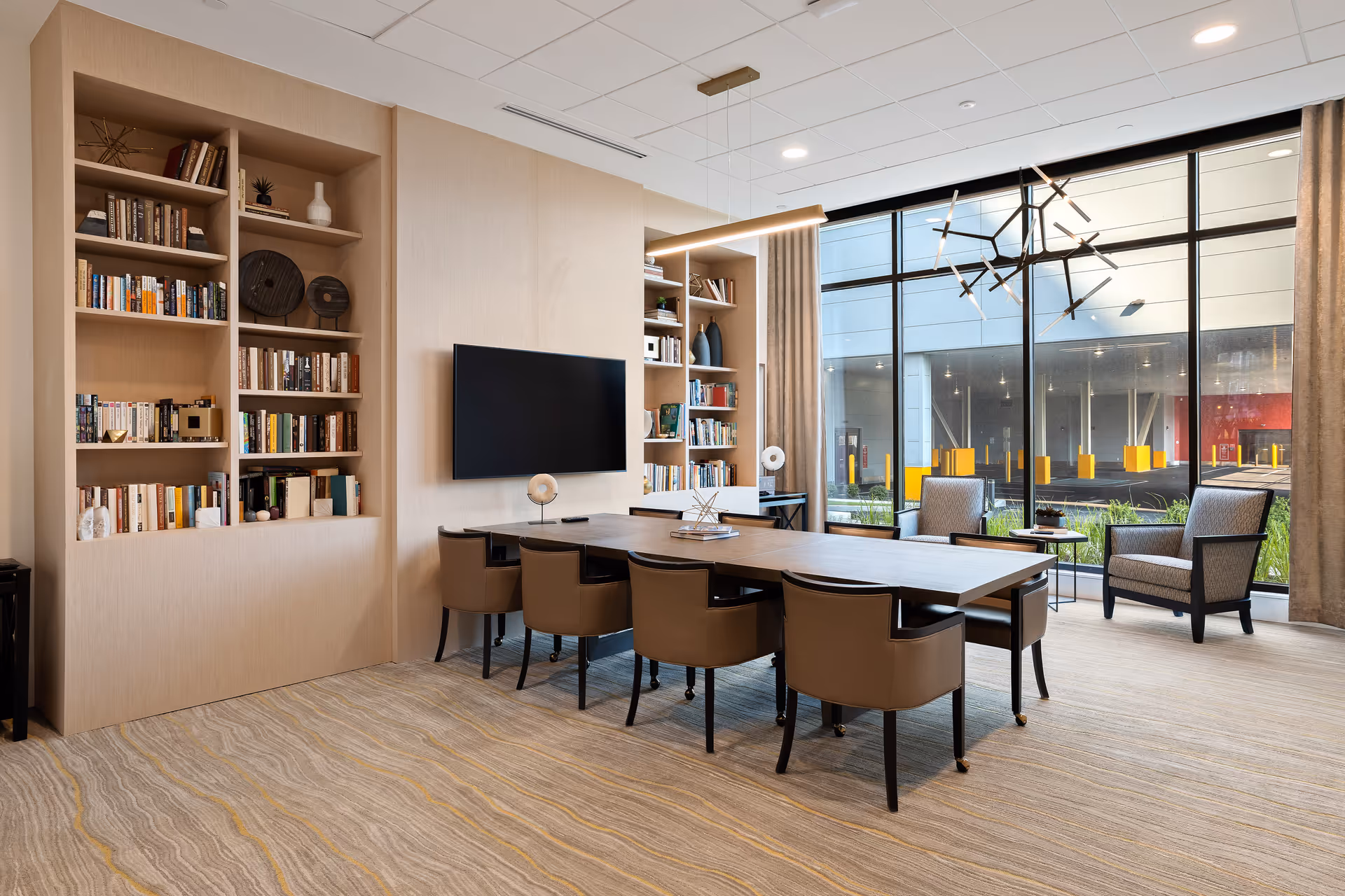 Bright communal room with a long wooden table and chairs, built-in bookshelves, a wall-mounted TV, and large floor-to-ceiling windows.