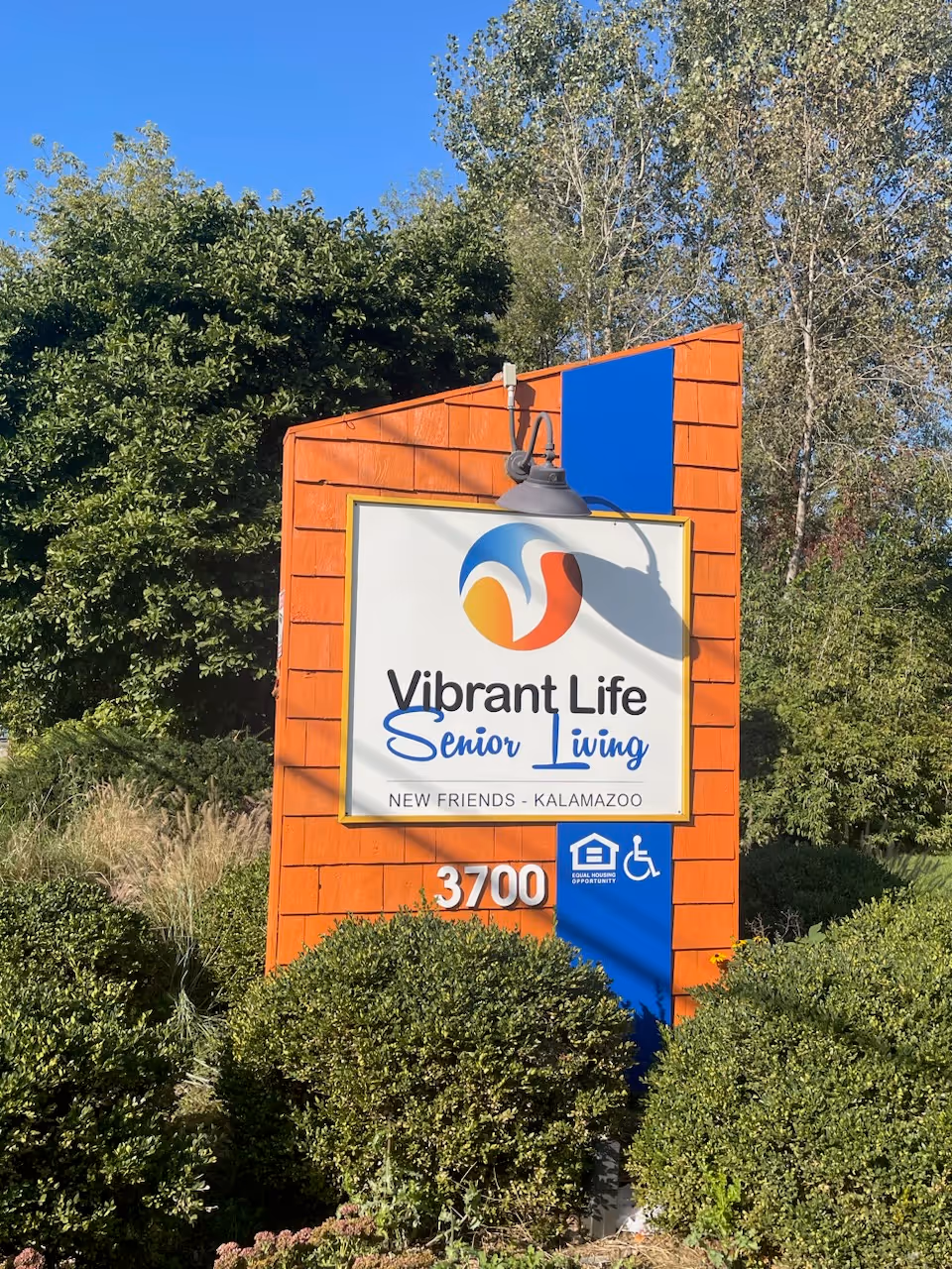 Orange building-front sign for Vibrant Life Senior Living Kalamazoo with greenery.