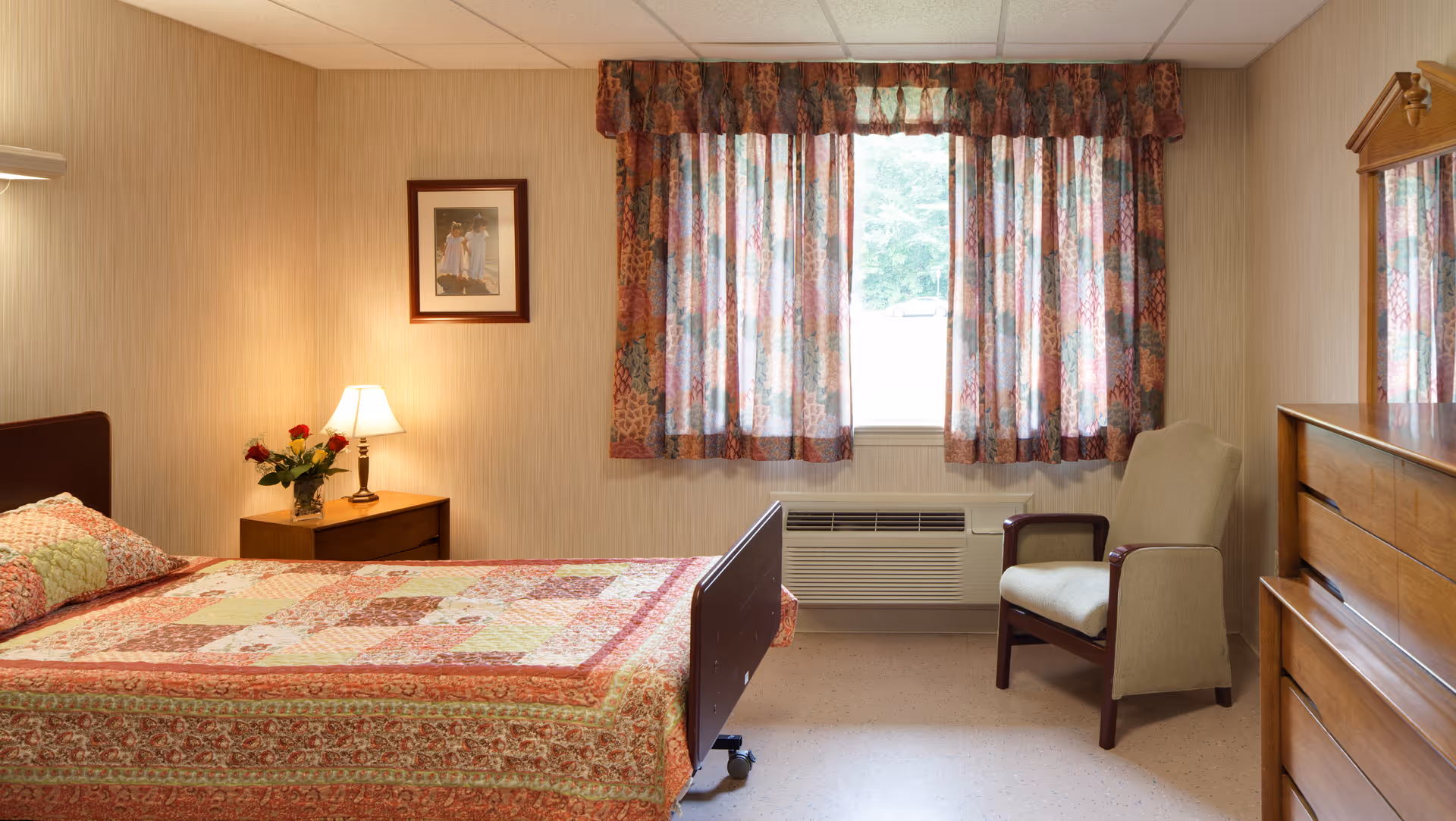 A cozy bedroom in a senior living facility featuring a bed with a patterned quilt, a wooden nightstand with a lamp and a vase of red roses, a framed picture on the wall, a window with floral curtains, an armchair, and a wooden dresser with a mirror.