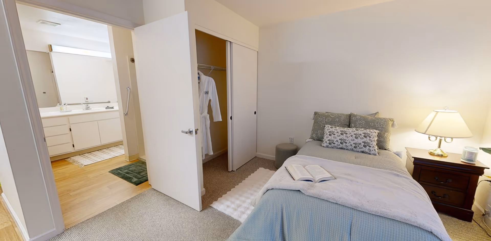 A cozy bedroom with a neatly made bed featuring green and floral pillows, a white blanket, and an open book on top. Next to the bed is a wooden nightstand with a lit lamp and a candle. The room has a closet with sliding doors, one of which is open showing a white robe hanging inside. The bedroom opens into a bathroom area with a large mirror, sink, and countertop visible.