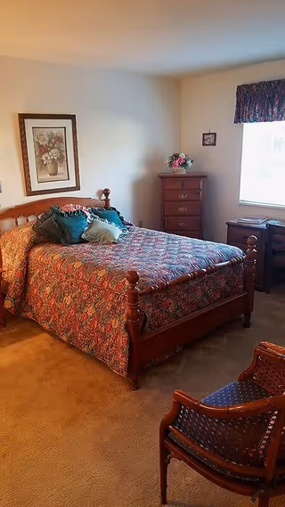 A cozy bedroom with a wooden bed frame and a floral patterned bedspread. The bed has several decorative pillows in green and beige. There is a wooden chest of drawers with a flower arrangement on top, a framed floral painting on the wall, a window with a floral valance, and a wooden chair with a blue cushion in the foreground.