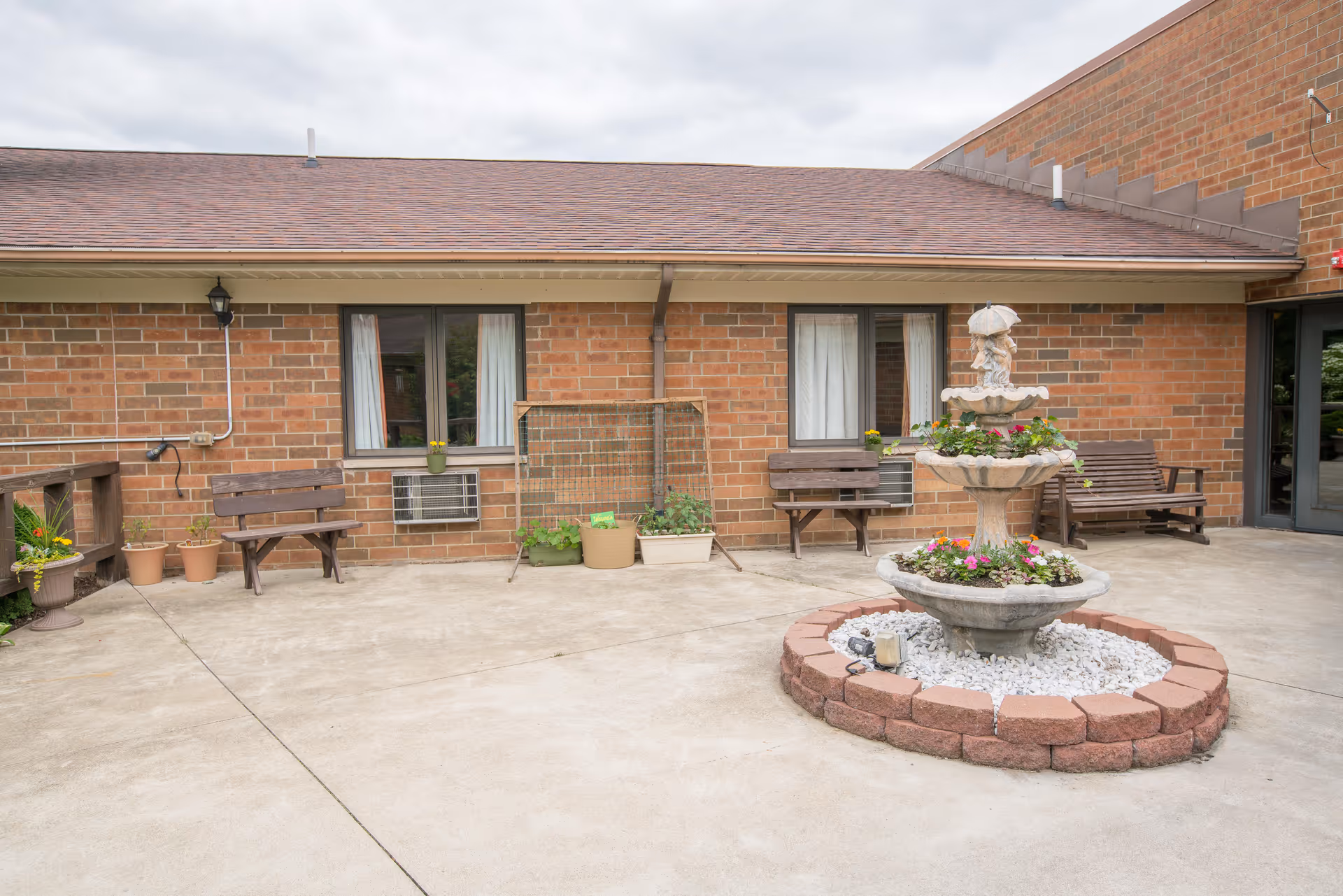 Outdoor courtyard area with a three-tiered stone fountain decorated with flowers in the center, surrounded by a circular brick border filled with white stones. The background shows a brick building with two windows, each with an air conditioning unit below, and three wooden benches placed against the wall. Several potted plants are arranged along the left side near a wooden railing. The sky is overcast.