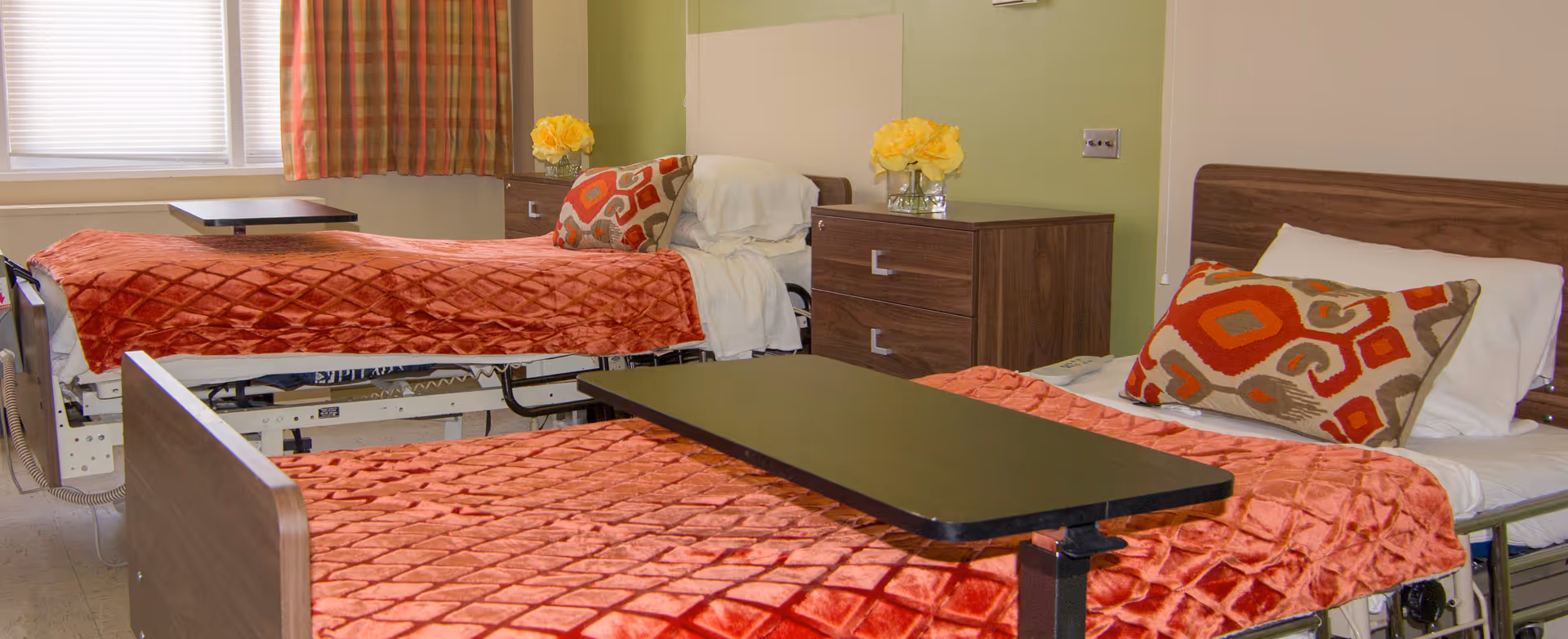Two hospital-style beds with red quilts, nightstands, and a central table in a shared patient room.