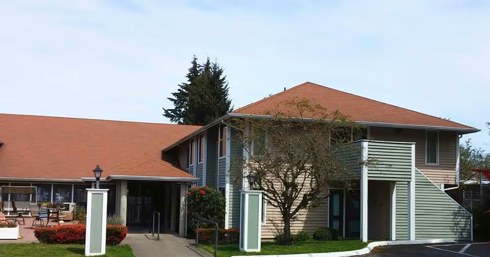 Exterior view of a two-story residential building with a red roof and beige siding, featuring an outdoor staircase and a small tree in front. There is a walkway leading to the entrance, surrounded by green grass and shrubs.
