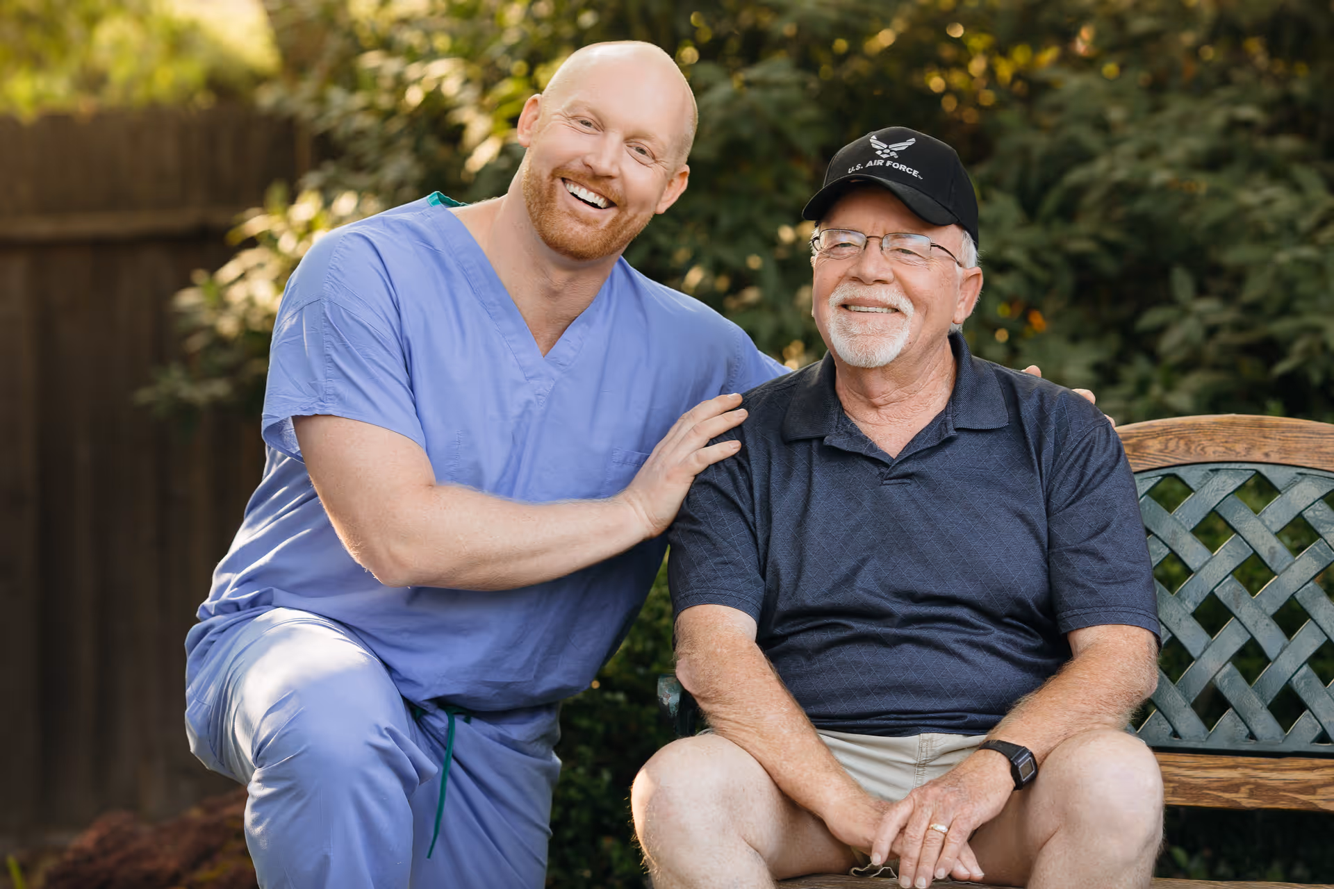 A caregiver in scrubs smiles beside an elderly man seated on a garden bench outdoors.
