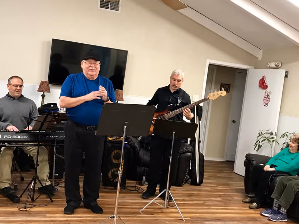 A group of three men performing music in a living room setting. One man is playing a keyboard, another is singing or speaking into a microphone, and the third is playing an electric bass guitar. Two women are seated on a couch watching the performance. The room has wooden flooring, a wall-mounted TV, and some decorative items on the walls and furniture.
