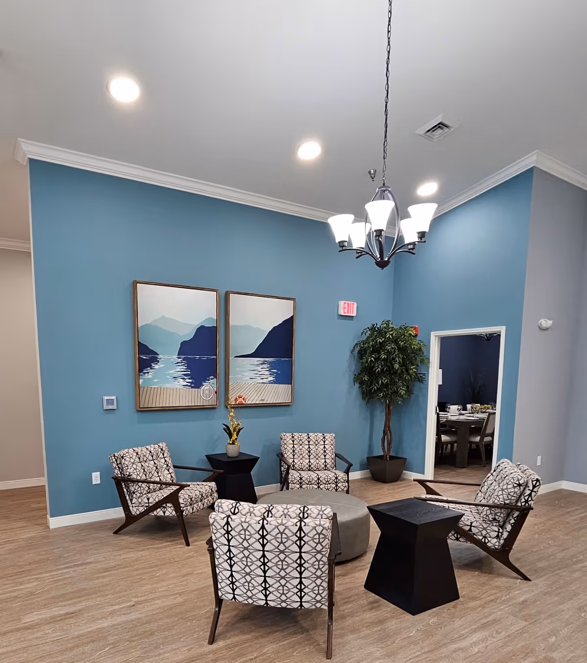 Blue-walled lounge with patterned chairs, round tables, and a chandelier in a senior living facility.
