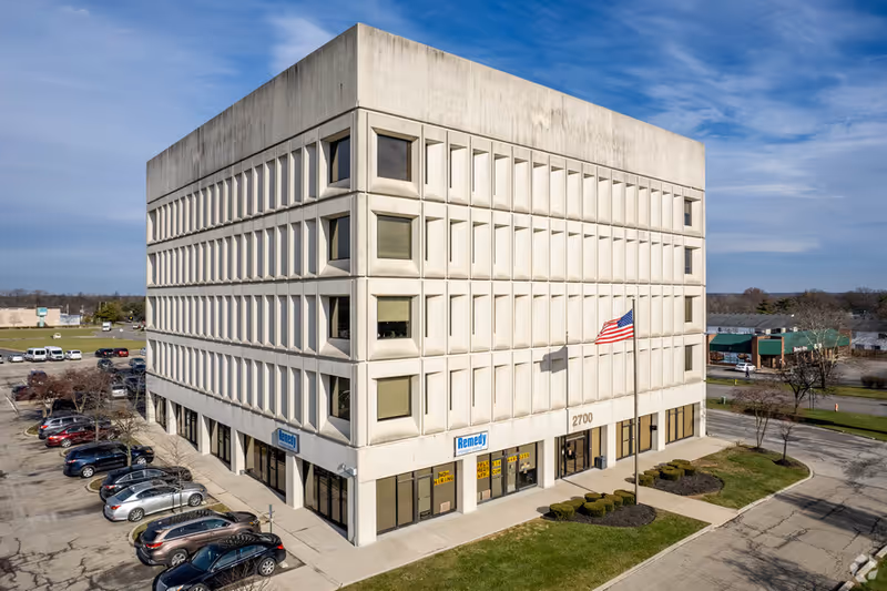 A modern beige, square, multi-story building with windows, parking, and an American flag.