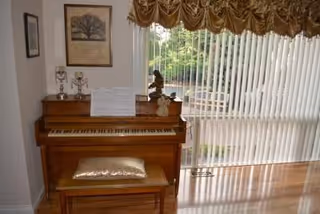 A wooden upright piano with bench sits beside a large window with vertical blinds.