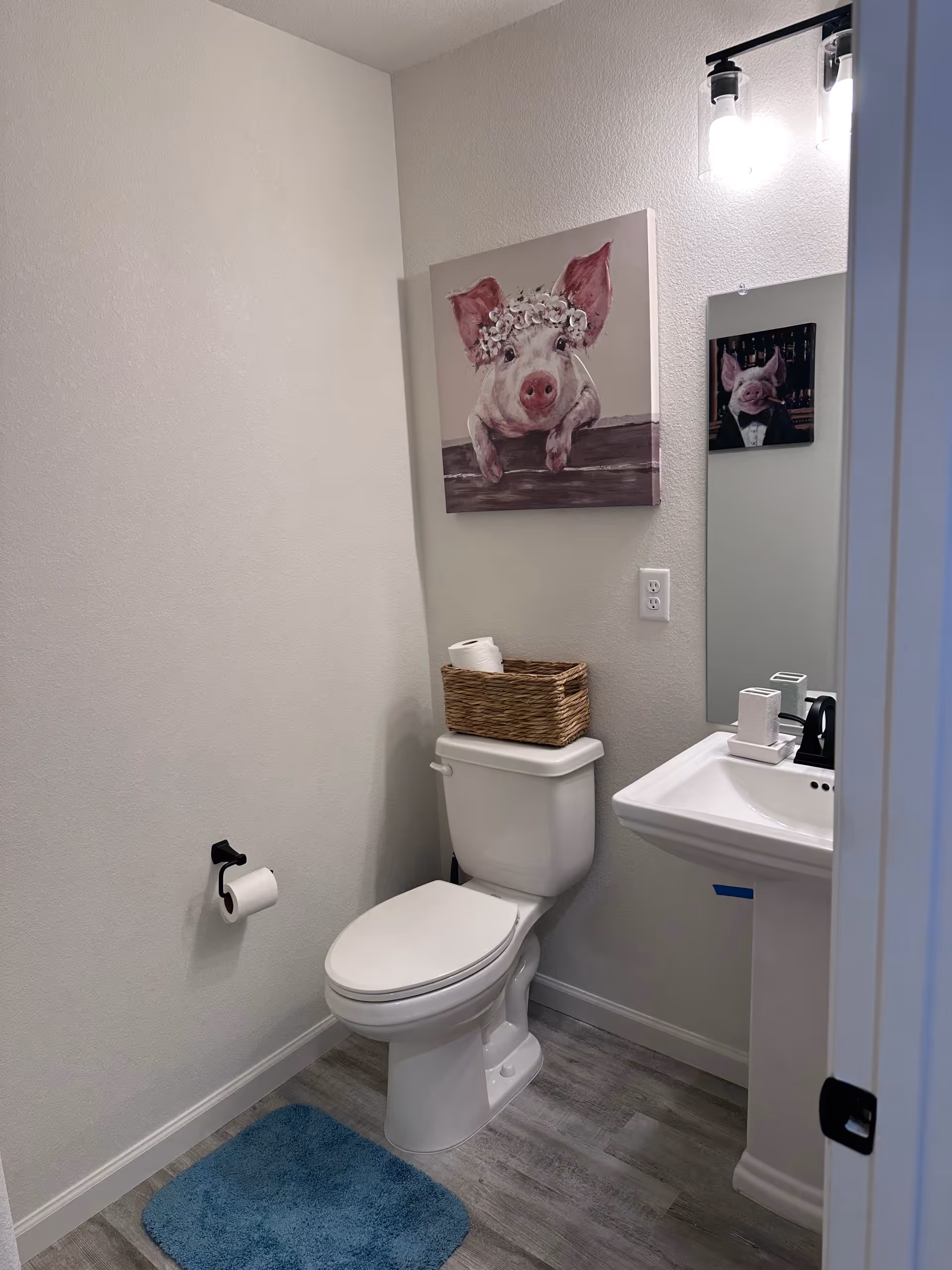 Small bathroom with toilet, sink, pig painting, wicker basket, and blue mat.
