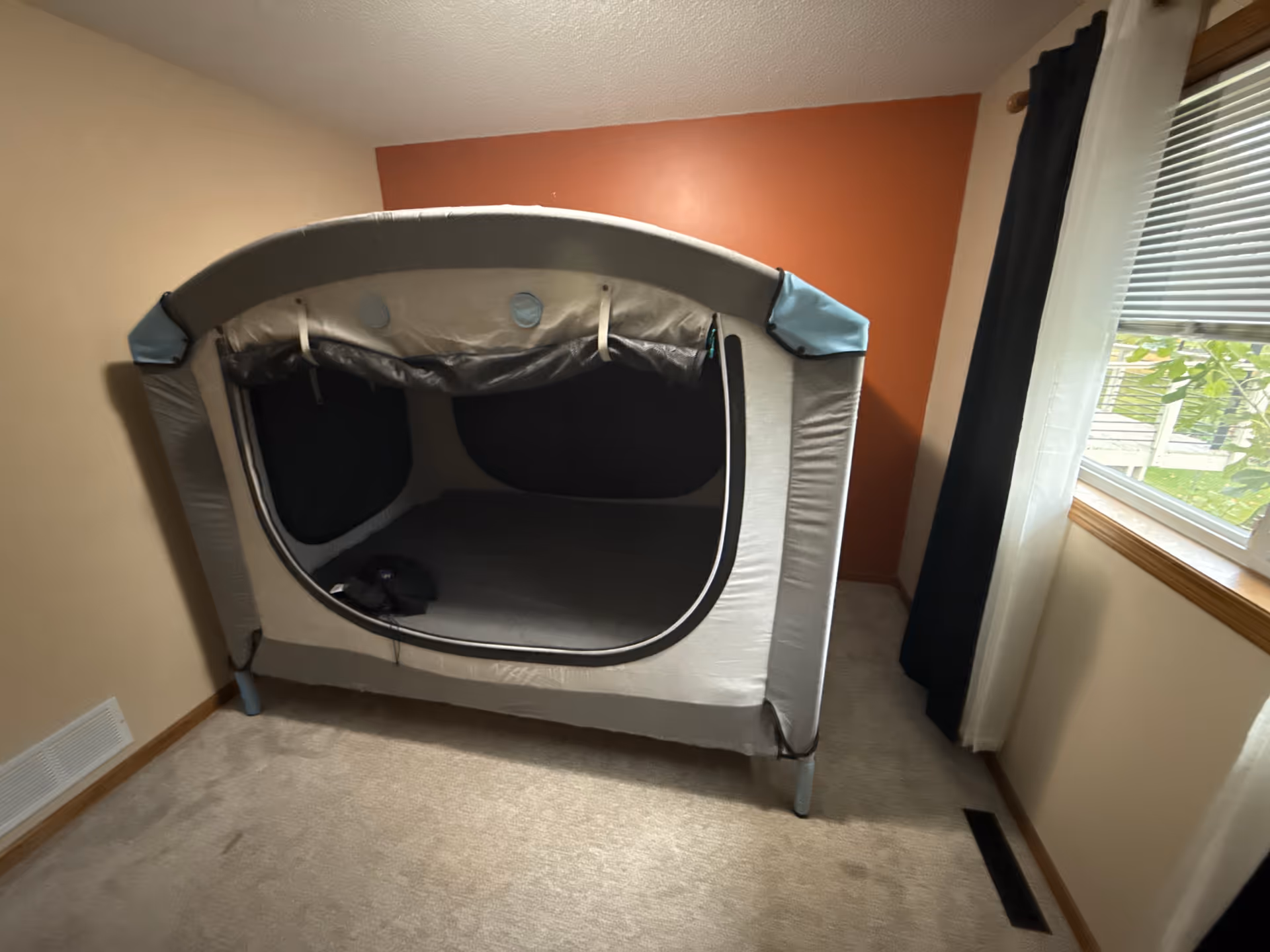 Grey tent-like structure inside a beige room with window, curtain; no people.