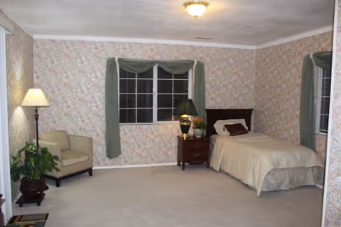 Cozy bedroom with floral wallpaper, bed, nightstand, lamps, window, and seating.