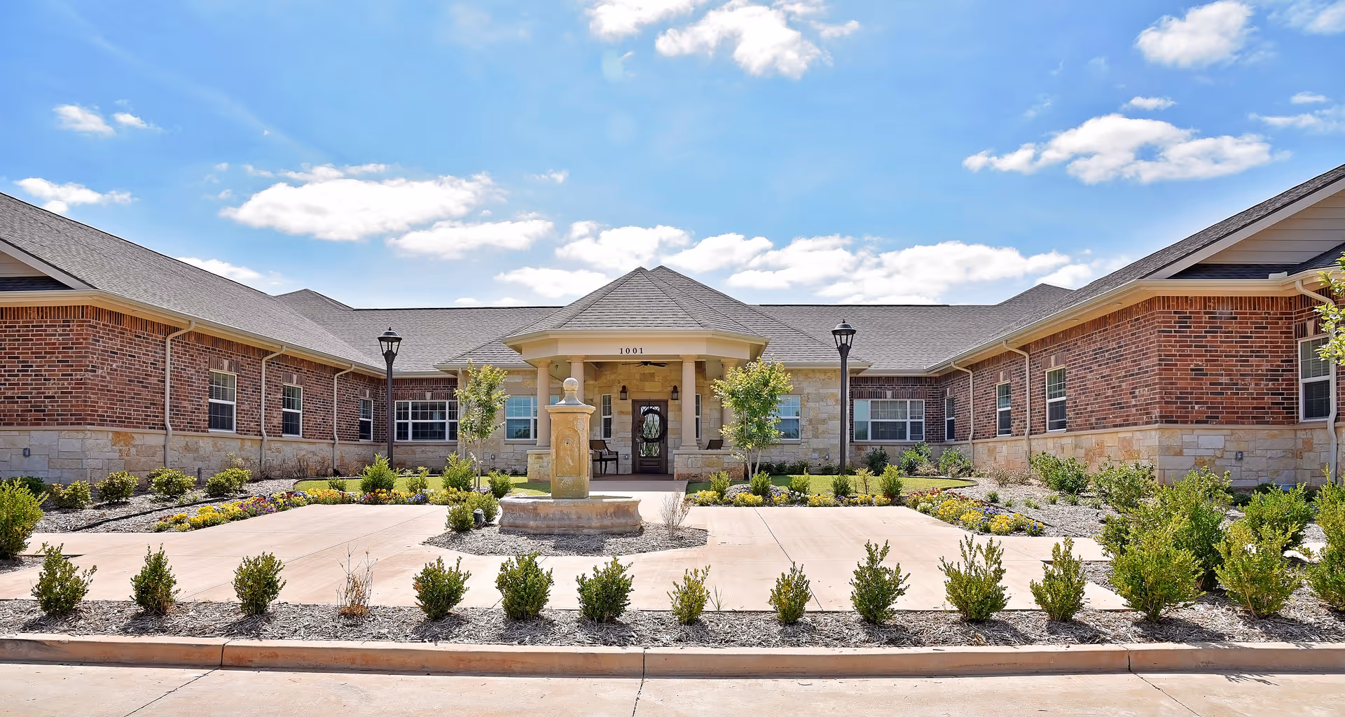 Front exterior of a single-story brick senior living facility with a central entrance, fountain, and landscaped driveway.