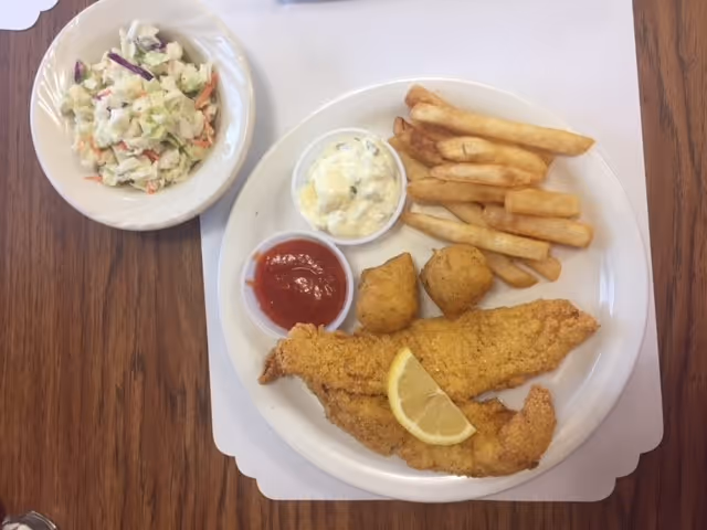 A meal consisting of fried fish fillets garnished with a lemon wedge, hush puppies, French fries, tartar sauce, and ketchup on a white plate. A side dish of coleslaw is served in a separate bowl on a wooden table.