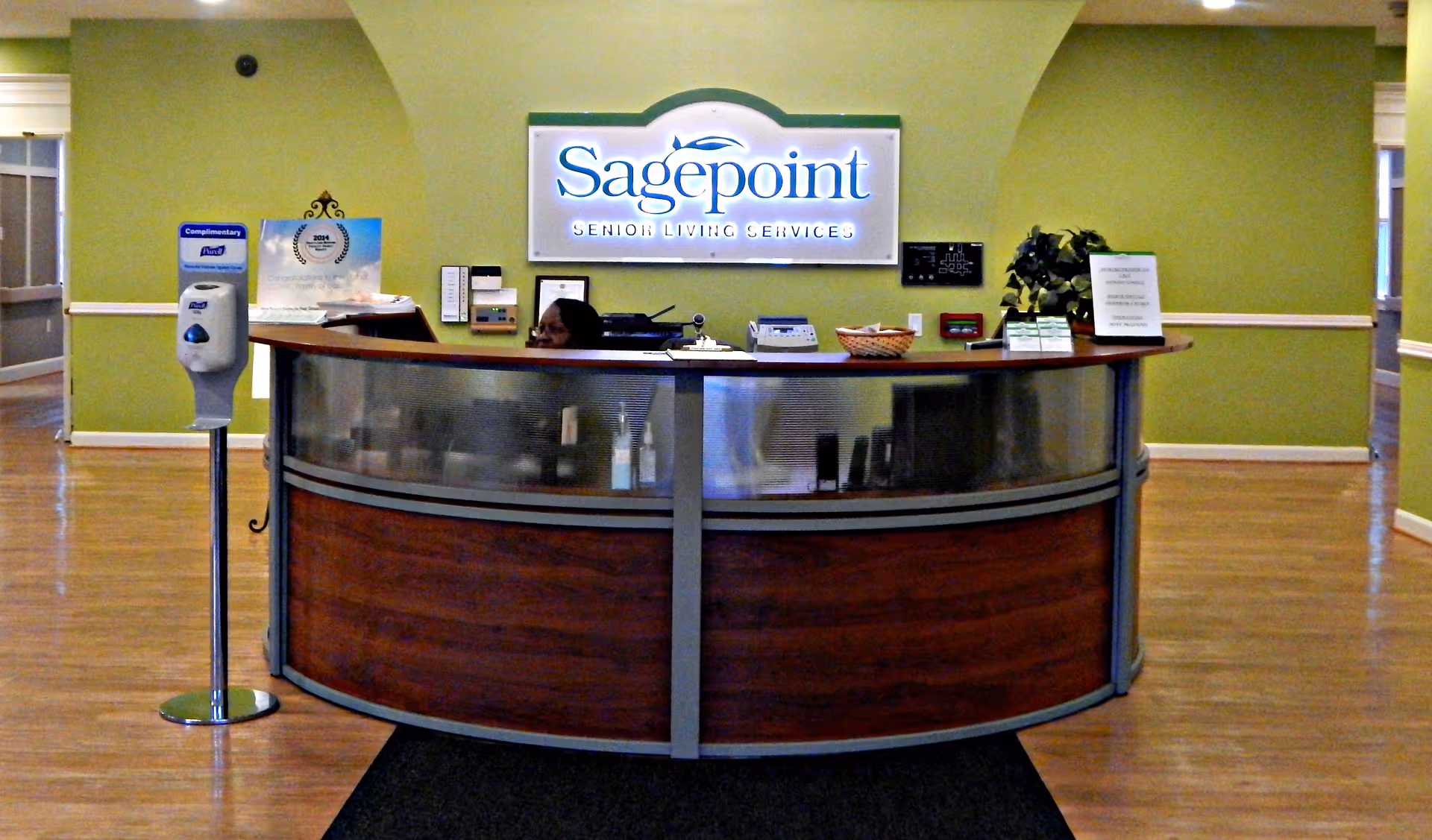 Curved wooden reception desk in Sagepoint Senior Living lobby, with receptionist behind it.