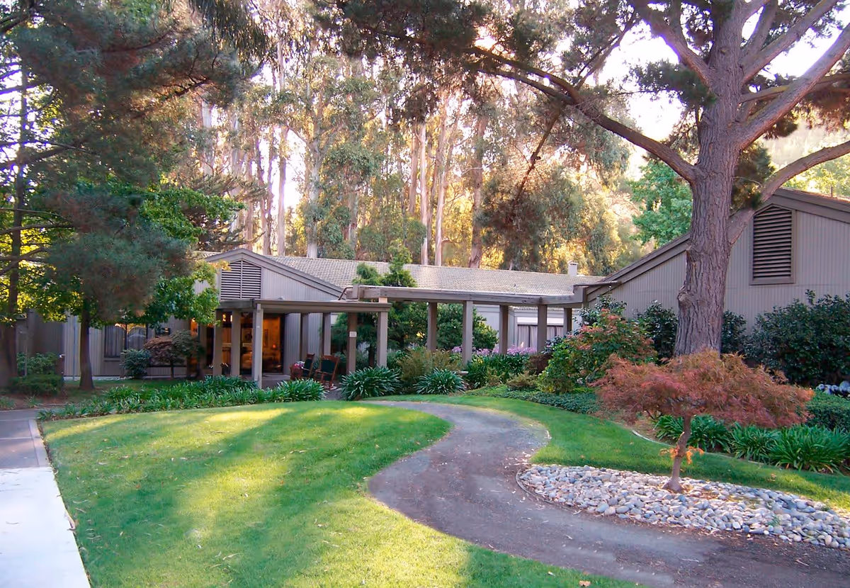 Front exterior of a single-story memory care building with a covered entrance, curved walkway, lawn, and surrounding trees.