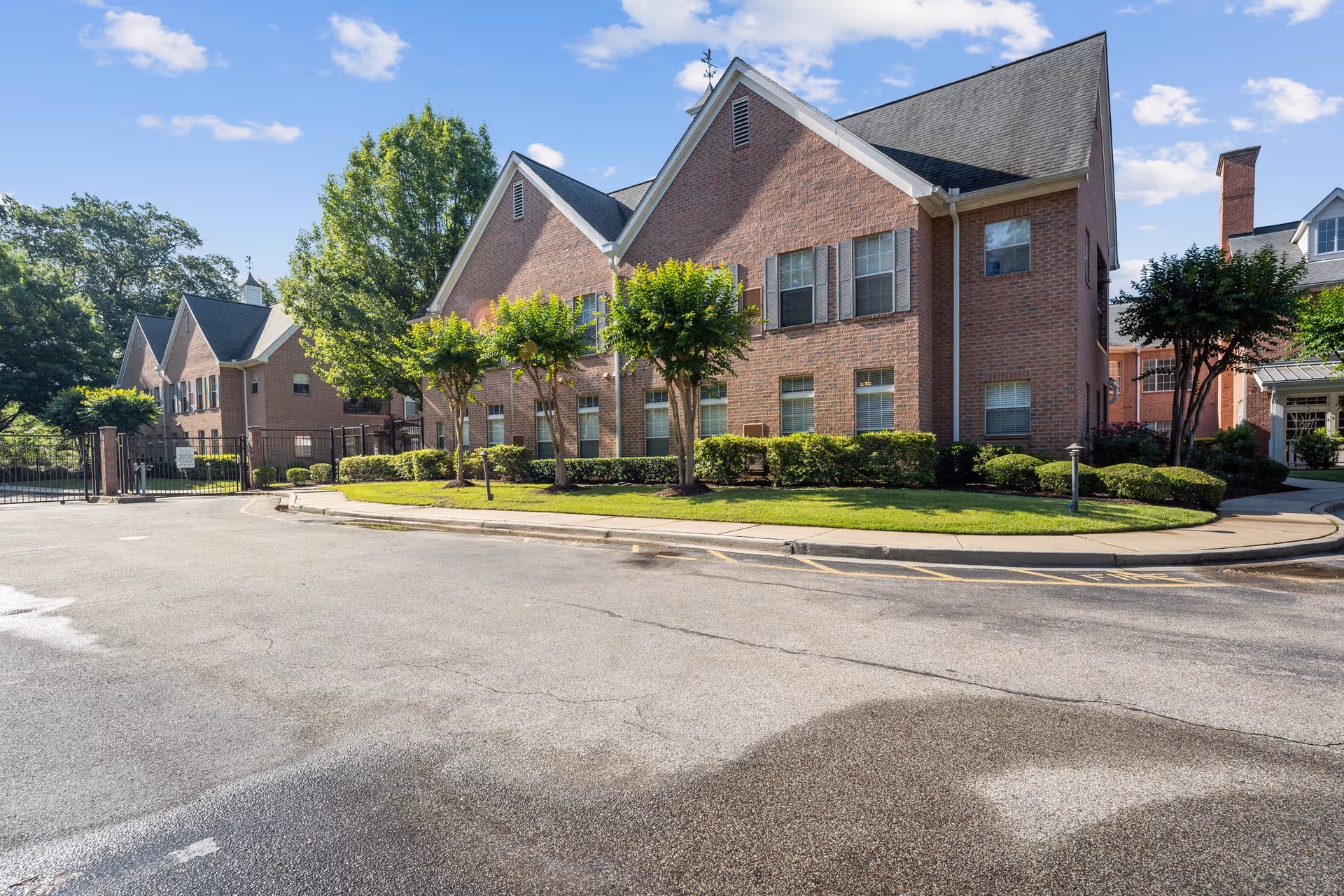 Brick two-story residential building with gabled roofs, trimmed shrubs and trees, and a paved driveway under a blue sky.