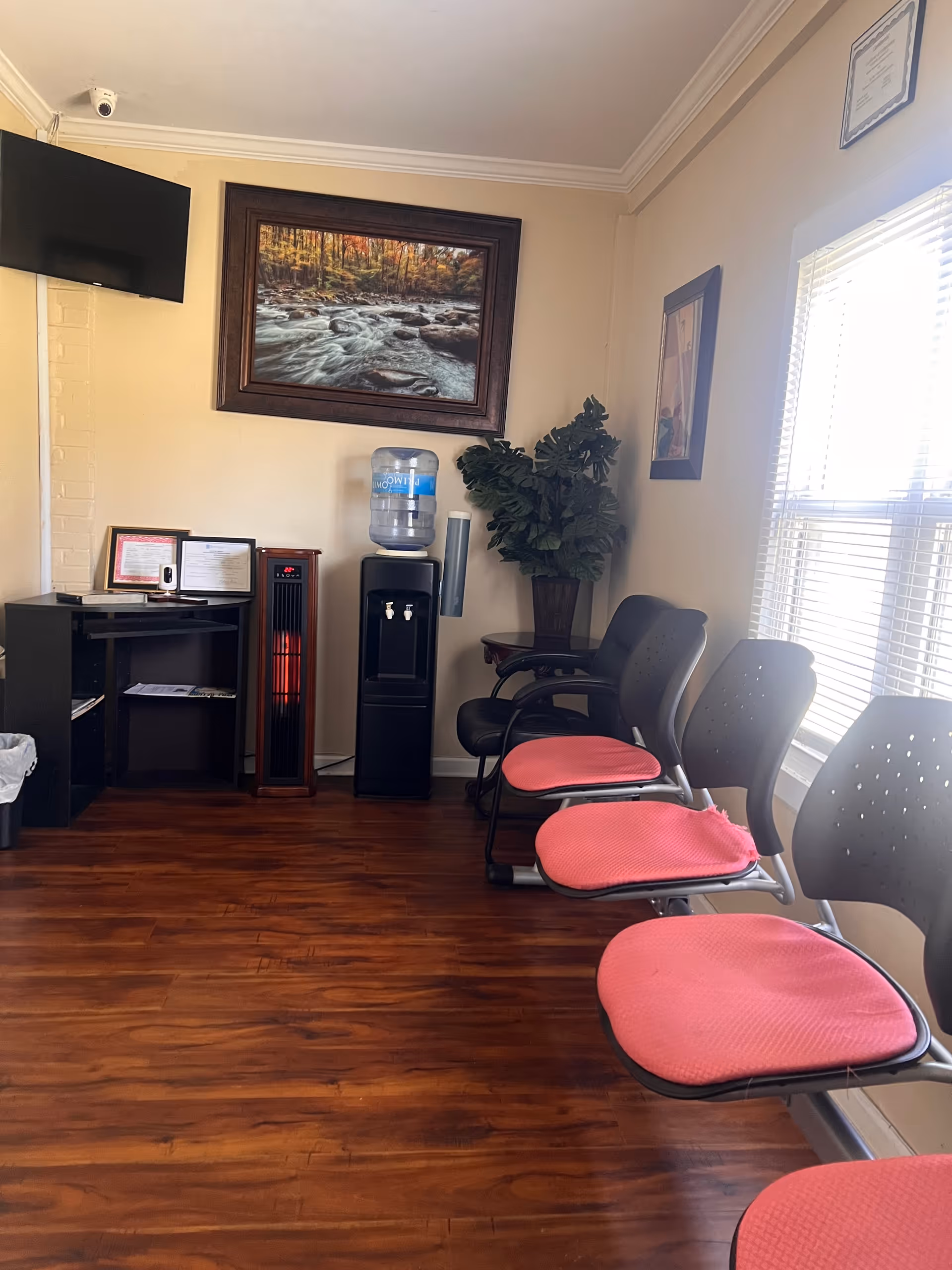 Interior waiting area with pink-cushioned chairs, a water cooler, framed landscape art, and window blinds.